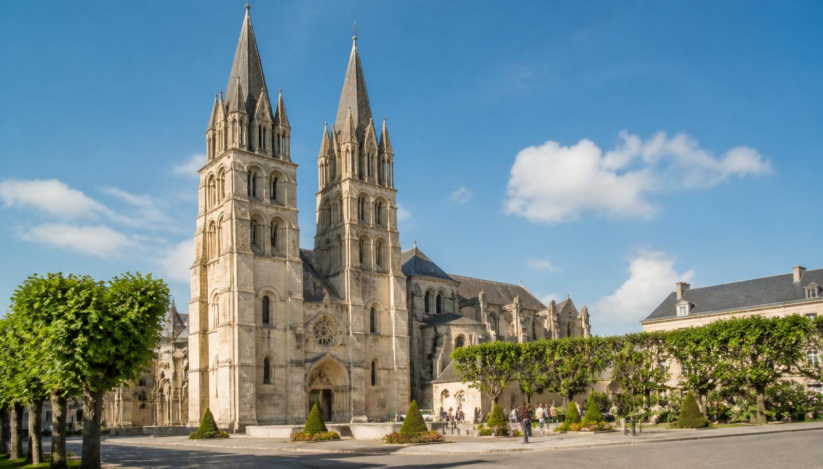 Illustration de l'abbaye aux Hommes à Caen : façade et flèches en pierre claire au-dessus d'une place plantée