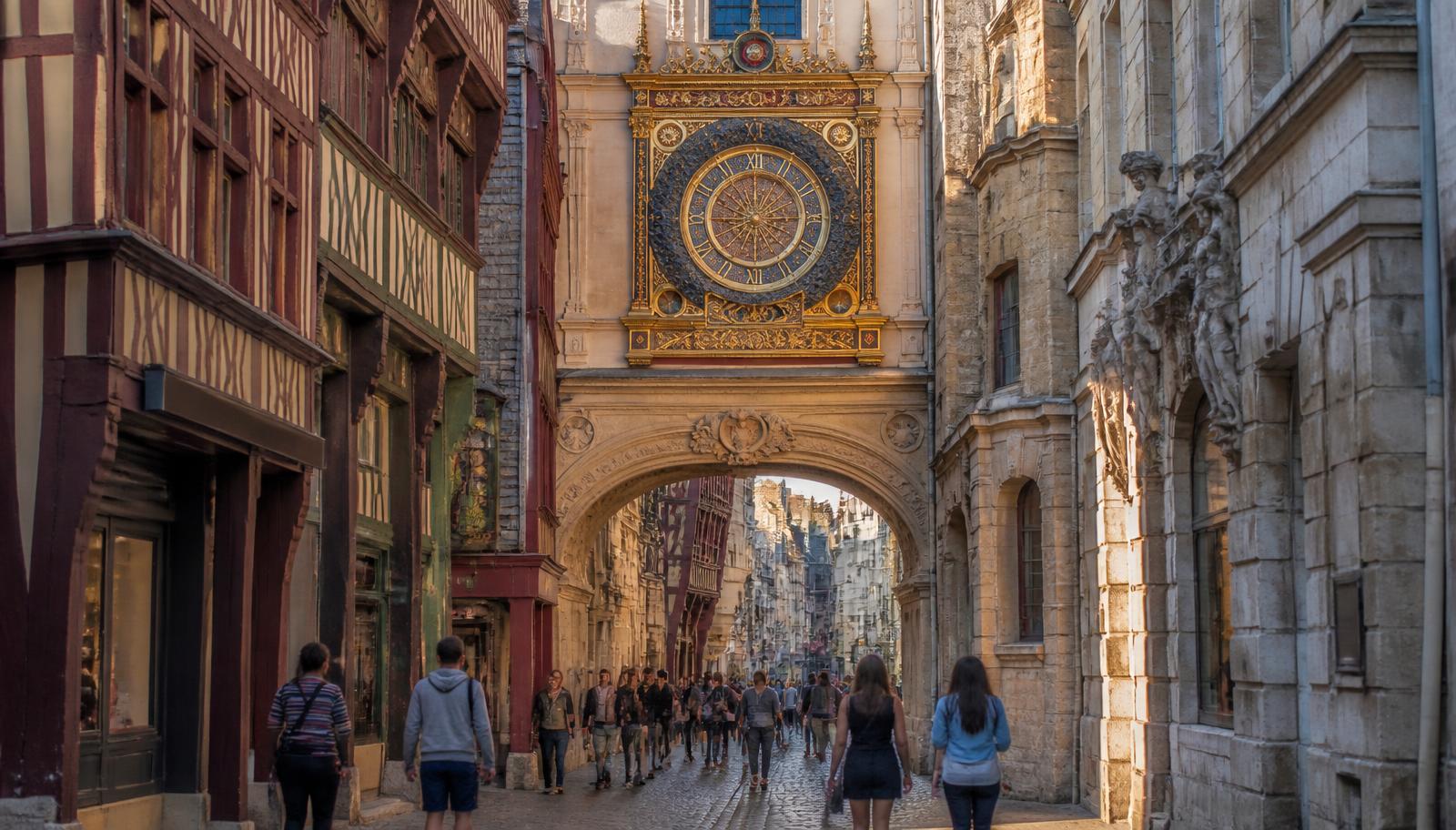 Illustration du Gros-Horloge de Rouen : arche renaissance et cadran doré au-dessus d'une rue pavée bordée de maisons à pans de bois